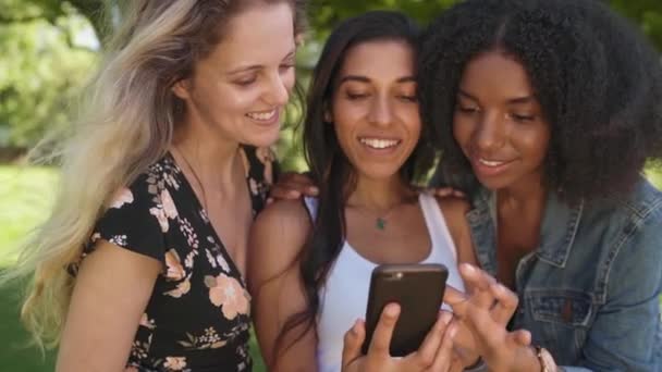 Веселые три различных подруги, зависающие в парке с помощью мобильного телефона и делящиеся цифровым контентом в социальных сетях, улыбающиеся и счастливые - глядя на приложение телефона — стоковое видео
