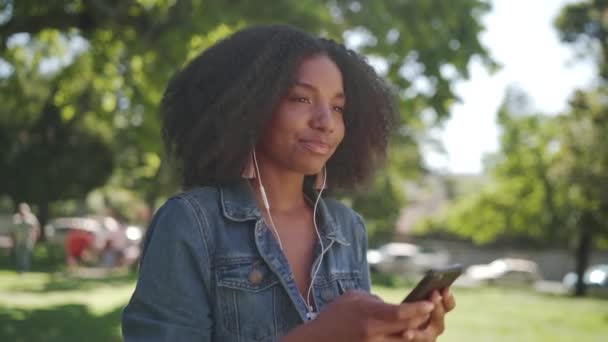 Egy afro-amerikai fiatal nő áll a parkban élvezi hallgatni a zenét a fülhallgató a mobiltelefonján - mosolygó és táncoló nő a parkban — Stock videók