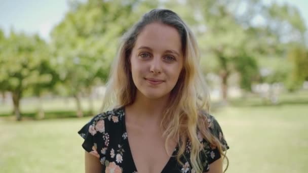 Porträtt av en attraktiv leende vågig blond ung kvinna tittar på kameran i parken - naturlig tonåring — Stockvideo