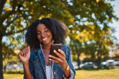 Afro-Amerikalı genç bir kadının portresi cep telefonuyla parkta müzik dinleyen, dans eden ve parkta eğlenen genç siyahi bir kadının kulaklıkla yaptığı müzik.