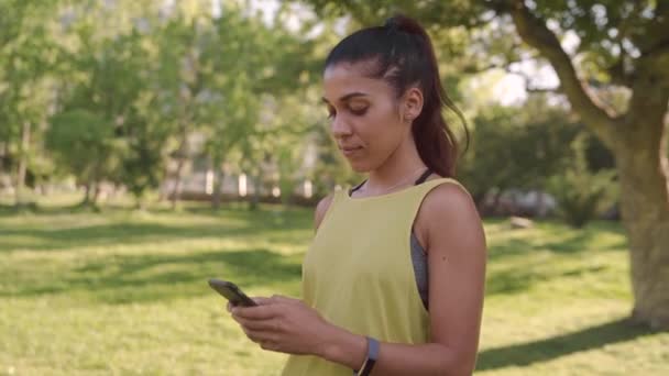 Retrato de um belo ajuste sorrindo jovem mulher em roupas esportivas de pé no parque mensagens de texto no telefone inteligente — Vídeo de Stock