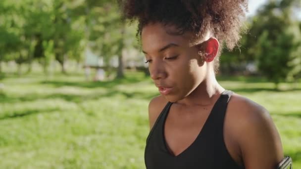 Una joven afroamericana de fitness que respira e inserta auriculares en sus oídos antes de correr por el parque — Vídeo de stock