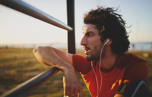 Vista lateral de un joven atleta agotado escuchando música en auriculares apoyados en bares en el parque del gimnasio mirando hacia el atardecer —  Fotos de Stock