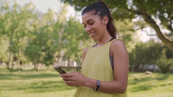 Dolly disparó a una joven en forma sonriente usando su teléfono móvil mirando hacia el parque — Vídeo de stock