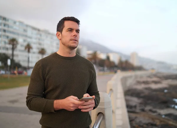 Retrato de um jovem bonito e pensativo em pé na rua da cidade segurando o telefone celular na mão olhando para longe — Fotografia de Stock