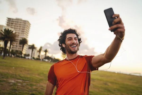 Retrato sonriente de un joven caucásico en forma con auriculares en sus oídos haciendo videollamadas desde un teléfono inteligente después de hacer ejercicio en el parque: un joven atleta masculino tomando una selfie — Foto de Stock