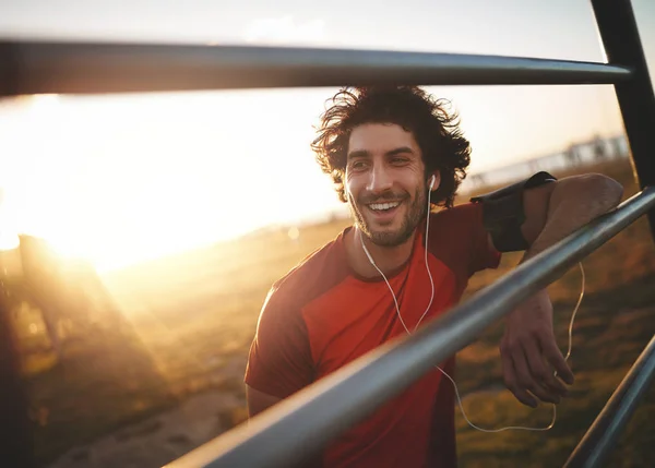 Retrato de un joven atleta sonriente con auriculares en las orejas apoyados en barras horizontales descansando después de correr —  Fotos de Stock