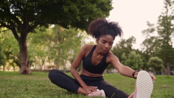 Egészséges, fiatal afro-amerikai nő fekete sportruházatban kinyújtóztatja a lábát, mielőtt fitnesz gyakorlatokat végezne a parkban. — Stock videók