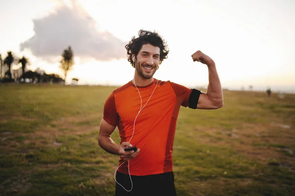 Retrato sonriente de un corredor masculino escuchando música en los auriculares y sosteniendo el teléfono móvil en la mano flexionando su músculo en el parque —  Fotos de Stock