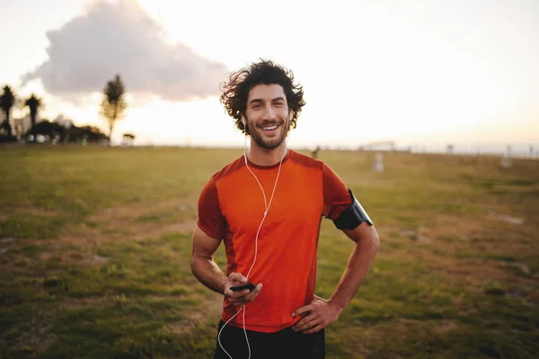 Retrato de un joven de fitness feliz con auriculares sosteniendo el teléfono móvil mientras está de pie en el parque mirando a la cámara —  Fotos de Stock