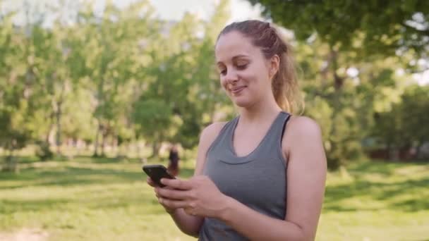 Caucasian fit young woman smiling while texting on mobile phone at the park in the morning on a sunny day — Stock Video