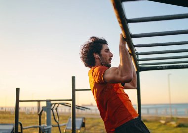 Kulaklıkları kulaklıklı, kaslı genç bir adamın parktaki spor salonunda barfiks çekerken yan görüntüsü.