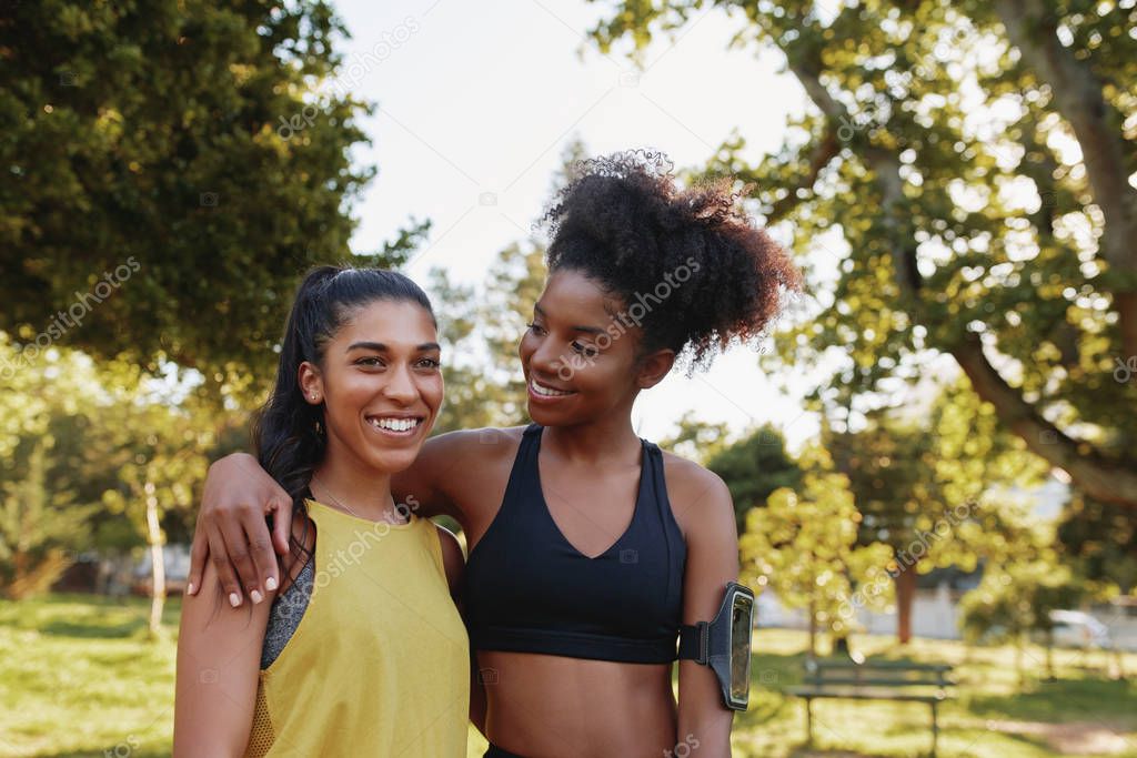 Smiling young women in sportswear standing arm in arm together at park - smiling multiethnic fitness friends in the park