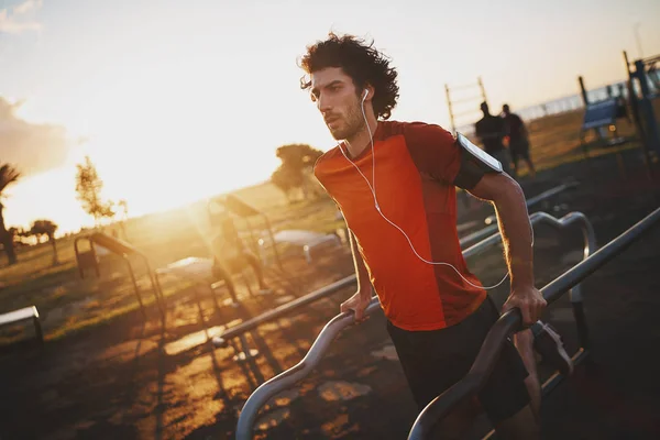 Ajuste joven escuchando música en los auriculares haciendo ejercicio en bares en el parque en el día soleado —  Fotos de Stock