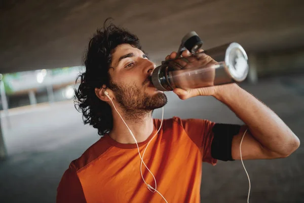 Close-up portrait of a handsome fit young man with earphones in his ears drinking water from a reusable water bottle — Stock Photo, Image