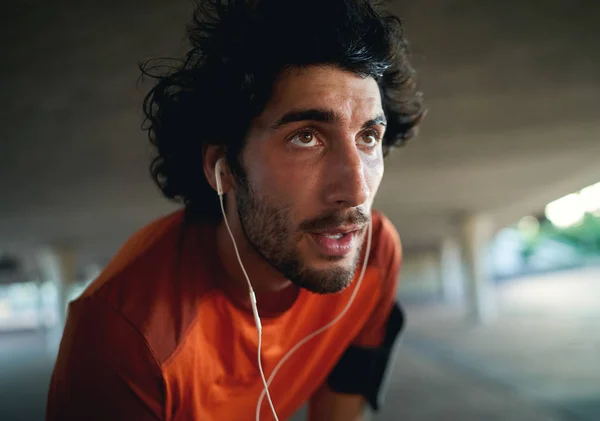 Retrato de un agotado corredor masculino serio y sudoroso con auriculares en los oídos relajándose después de correr mirando a la distancia —  Fotos de Stock