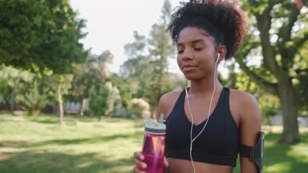 Porträtt av en glad afrikansk amerikansk idrottare ung kvinna lyssnar på musik på hörlurar dricka vatten från återanvändbar flaska i parken — Stockvideo