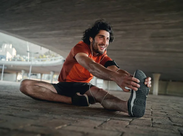 Retrato de un joven atleta sonriente con auriculares sentado debajo del puente de la ciudad estirando las piernas antes de trotar —  Fotos de Stock