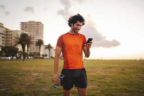 Веселий портрет спортивного молодого чоловіка, який дивиться на екран своїх смартфонів і слухає музику в своїх білих навушниках, стоячи в парку — стокове фото