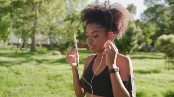 Atleta feliz mujer afroamericana corredora poniendo auriculares en sus oídos escuchando música en el teléfono inteligente en el brazalete en el parque — Vídeo de stock