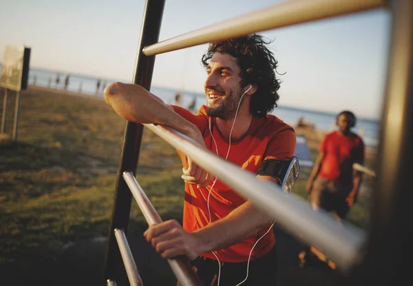 Portræt af en smilende ung atletisk mand nyder musikken fra hans øretelefoner hviler efter træning på udendørs gym - Stock-foto