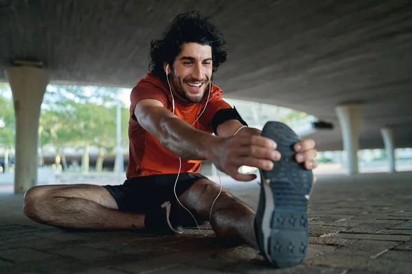 Retrato de un atleta feliz disfrutando de la música en sus auriculares calentándose y estirando sus piernas antes de trotar bajo el puente —  Fotos de Stock