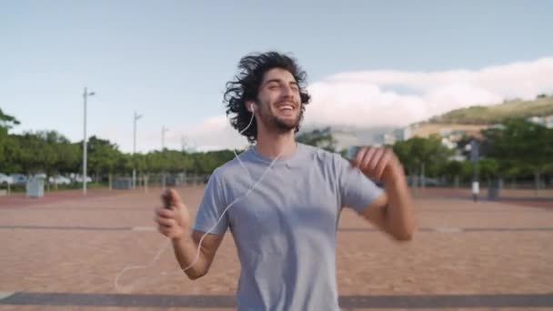 Молодий чоловік ходить і танцює до камери насолоджуючись і слухаючи музику на смартфоні через навушники в парку — стокове відео