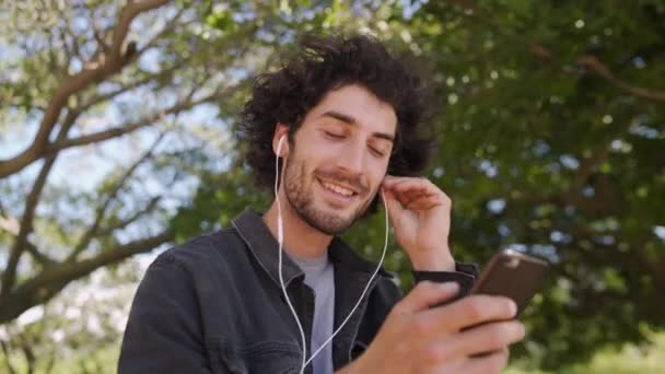 Портрет улыбающегося молодого человека с наушниками в ушах, болтающего в социальных сетях на смартфоне в парке — стоковое видео