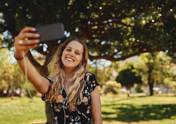 Retrato de una joven sonriente con auriculares en los oídos tomando una selfie en el teléfono móvil en el parque - mujer joven feliz — Foto de Stock