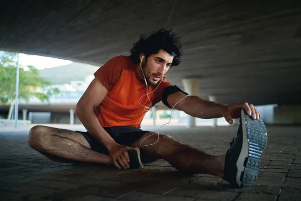 Joven atleta con auriculares en los oídos calentándose y estirando las piernas antes de correr en la calle de la ciudad bajo el puente —  Fotos de Stock