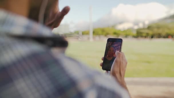 Por encima de la toma de hombro de hombre charlando en línea - haciendo videollamada en su teléfono inteligente con su amigo en el parque — Vídeo de stock