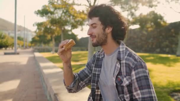 Portret van een jongeman die op zonnige dag in de frisse lucht in het park boterham eet — Stockvideo