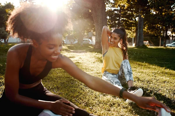 Due giovani amiche diverse sedute sul prato alla luce del sole che fanno esercizio di stretching nel parco - amici che si scaldano prima di fare esercizio — Foto Stock
