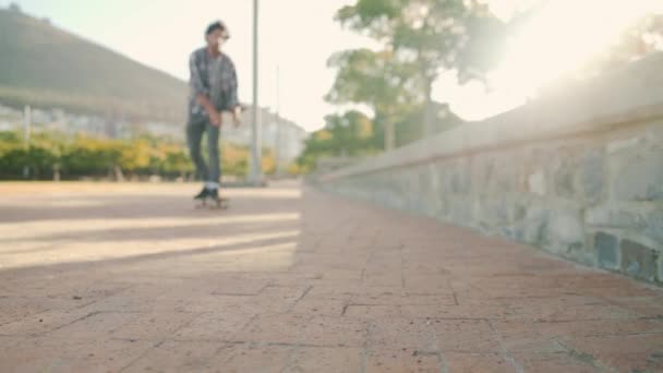 Primer plano de los pies de un hombre joven después de montar en monopatín hacia la cámara sentado en una pared en el parque en un día soleado con destello de sol — Vídeos de Stock