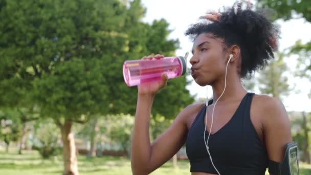 Αφροαμερικάνα αθλήτρια Νεαρή γυναίκα πίνει νερό από επαναχρησιμοποιήσιμο μπουκάλι και αναπνέει βαριά μετά την πρωινή προπόνηση στο πάρκο — Αρχείο Βίντεο