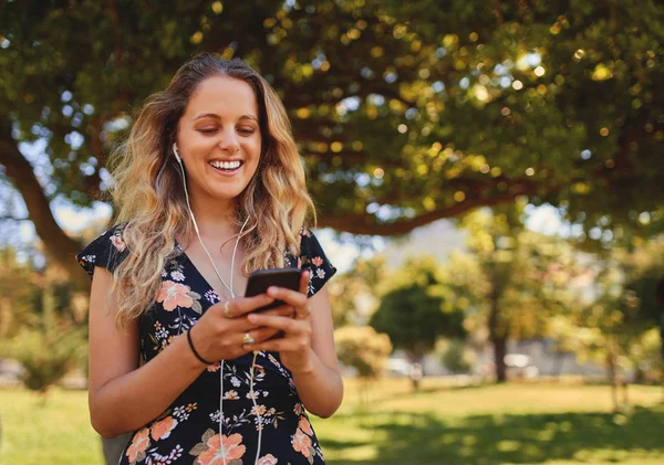 Retrato de uma jovem loira sorridente feliz com fones de ouvido nos ouvidos mensagens de texto em seu telefone inteligente no parque em um dia ensolarado — Fotografia de Stock