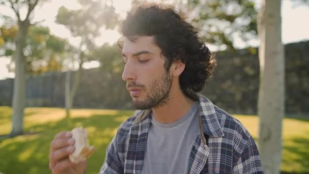 Guapo joven sonriente sentado en el parque en un día soleado disfrutando de comer sándwich - almorzando en el parque en un día soleado de verano — Vídeo de stock