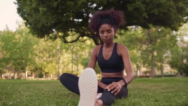 Retrato de una joven afroamericana sentada sobre hierba verde estirando las piernas en el parque antes de correr — Vídeos de Stock