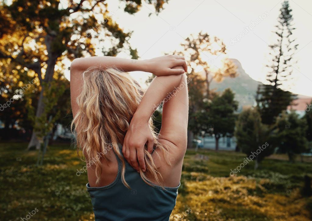 Rear view of blonde young woman in sportswear stretching her arm in the park - sporty woman stretching in the park 