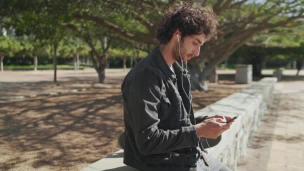 Retrato de un joven de la cadera sonriente patinador feliz con auriculares en los oídos mensajes de texto en el teléfono móvil en el parque — Vídeo de stock