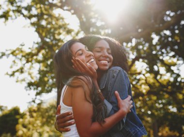 Smiling portrait of a diverse young female with eyes closed cuddling and loving each other in the park with sunlight shining through