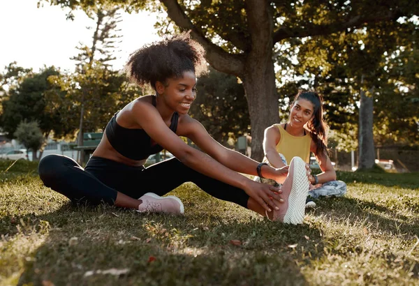 Sonriendo a dos amigas jóvenes en forma deportiva que estiran sus piernas en el parque: una negra afroamericana que se calienta con su amiga antes de hacer ejercicio en el parque rodeada de árboles — Foto de Stock