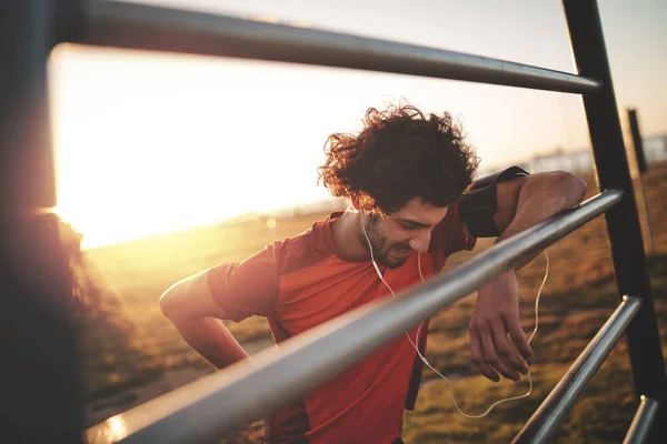 Bello giovane corridore maschio che ascolta musica su auricolari a riposo dopo aver fatto jogging al parco in una giornata di sole — Foto Stock