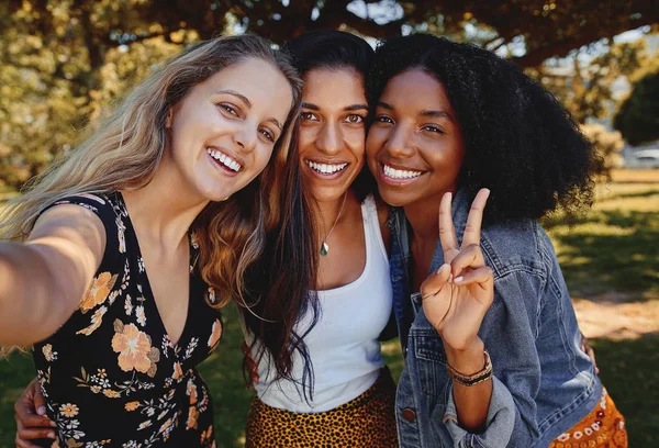 Retrato de três sorridentes amigas multiétnicas felizes tirando uma selfie no parque em um dia ensolarado — Fotografia de Stock