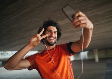 Portrait of a smiling young and handsome man with mobile smartphone pulling a peace sign and taking a selfie 
