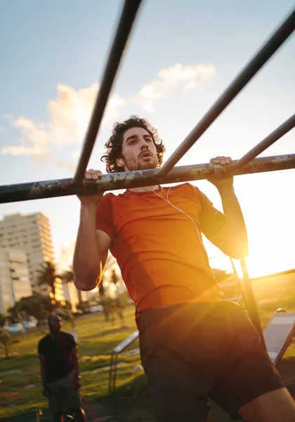 Ajuste o jovem com fones de ouvido em seus ouvidos treinando seus músculos dos braços em bares no ginásio ao ar livre no verão homem fazendo pullups ao ar livre — Fotografia de Stock