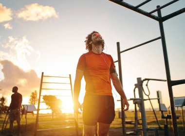 Happy fit young man enjoying listening to music on earphones after exercising in the outdoor gym park on summer sunny day