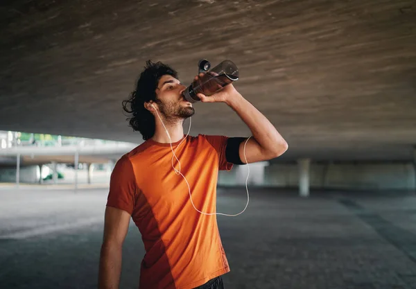Young male athlete with earphone in his ears drinking water from reusable bottle standing under the concrete bridge