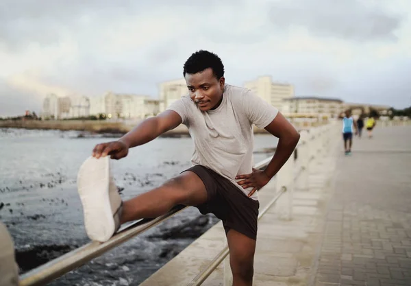 Hombre decidido a doblar para tocar los dedos de los pies mientras pone la pierna en la barandilla mientras hace ejercicio cerca de la playa por la mañana —  Fotos de Stock