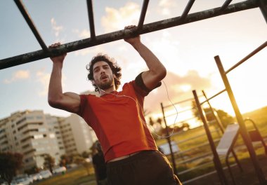 Portrait of a young muscular man listening to music on earphones working out on monkey bars doing pull-ups in outdoor gym